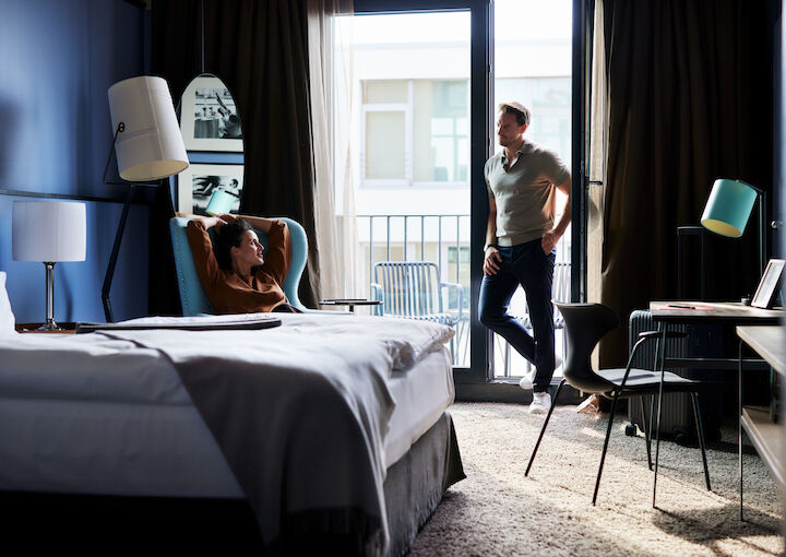 Woman relaxes in armchair, man leans by balcony door in M-Room